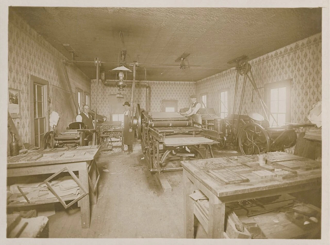 Photograph of a printing office in Goldthwaite with several type setting stations visible throughout the space and a large printing press in the center of the room. Three men stand in the back of the room, the one on the left wearing a dark waistcoat and light colored shirt and the two on the right wearing hats and aprons over their attire. A typed note is adhered to the bottom edge of the photo card that reads, "Goldthwaite Eagle about 1910. R. M. Thompson & printers. Notice no electric lights or leno type." The same message is written in pen underneath the label.